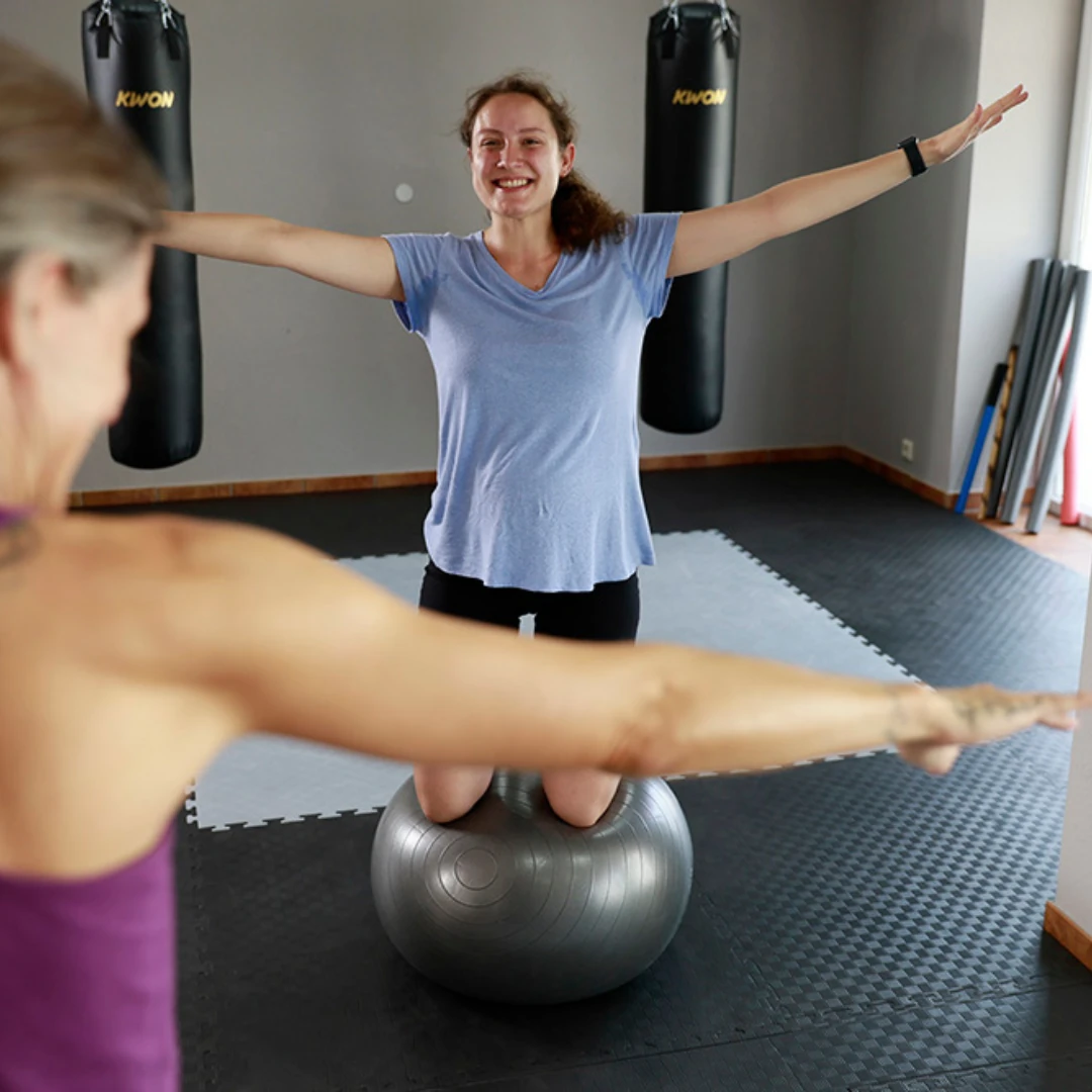 Eine Frau steht mit ausgebreiteten Armen auf einem Gymnastikball in einem Fitnessstudio, während eine andere Person im Vordergrund trainiert.