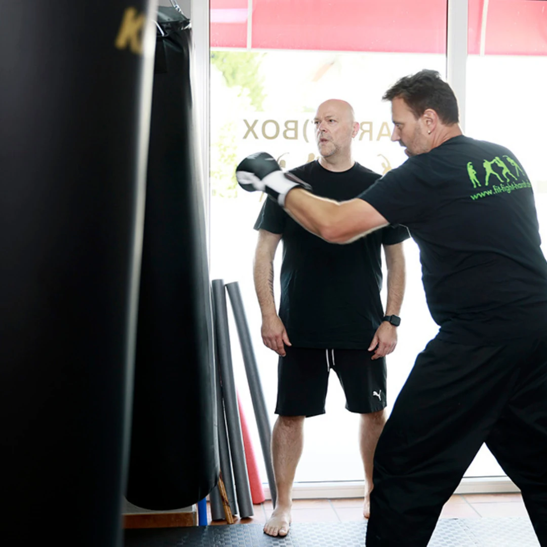 Zwei Männer trainieren im Fitnessstudio, einer trägt Boxhandschuhe und führt einen Schlag aus, während der andere daneben steht.