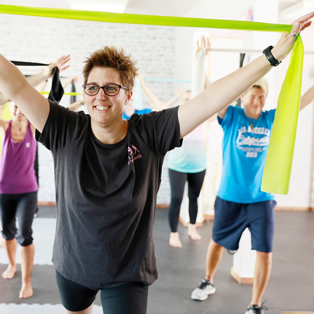 Eine Gruppe von Menschen führt in einem Fitnessstudio Übungen mit Gummibändern durch, wobei eine Person im Vordergrund lächelnd die Arme ausgestreckt hält.