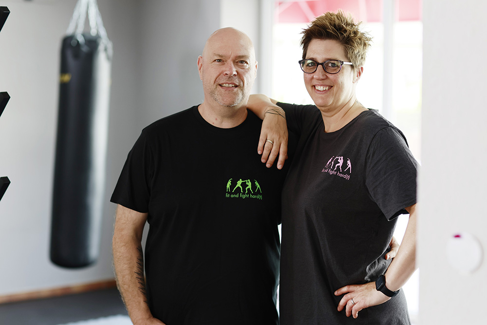 Zwei Personen stehen lächelnd nebeneinander in einem Fitnessstudio, wobei eine Person den Arm um die andere legt.